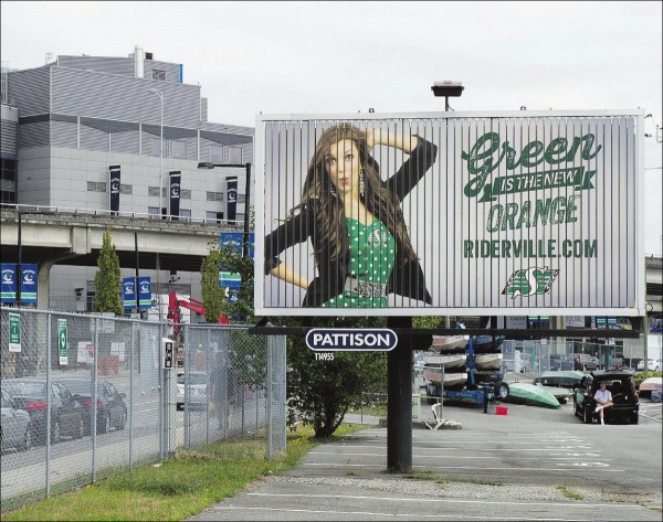 A Pattison billboard shows a white woman wearing a green Saskatchewan RoughRiders shirt. The text on the billboard reads Green is the New Orange