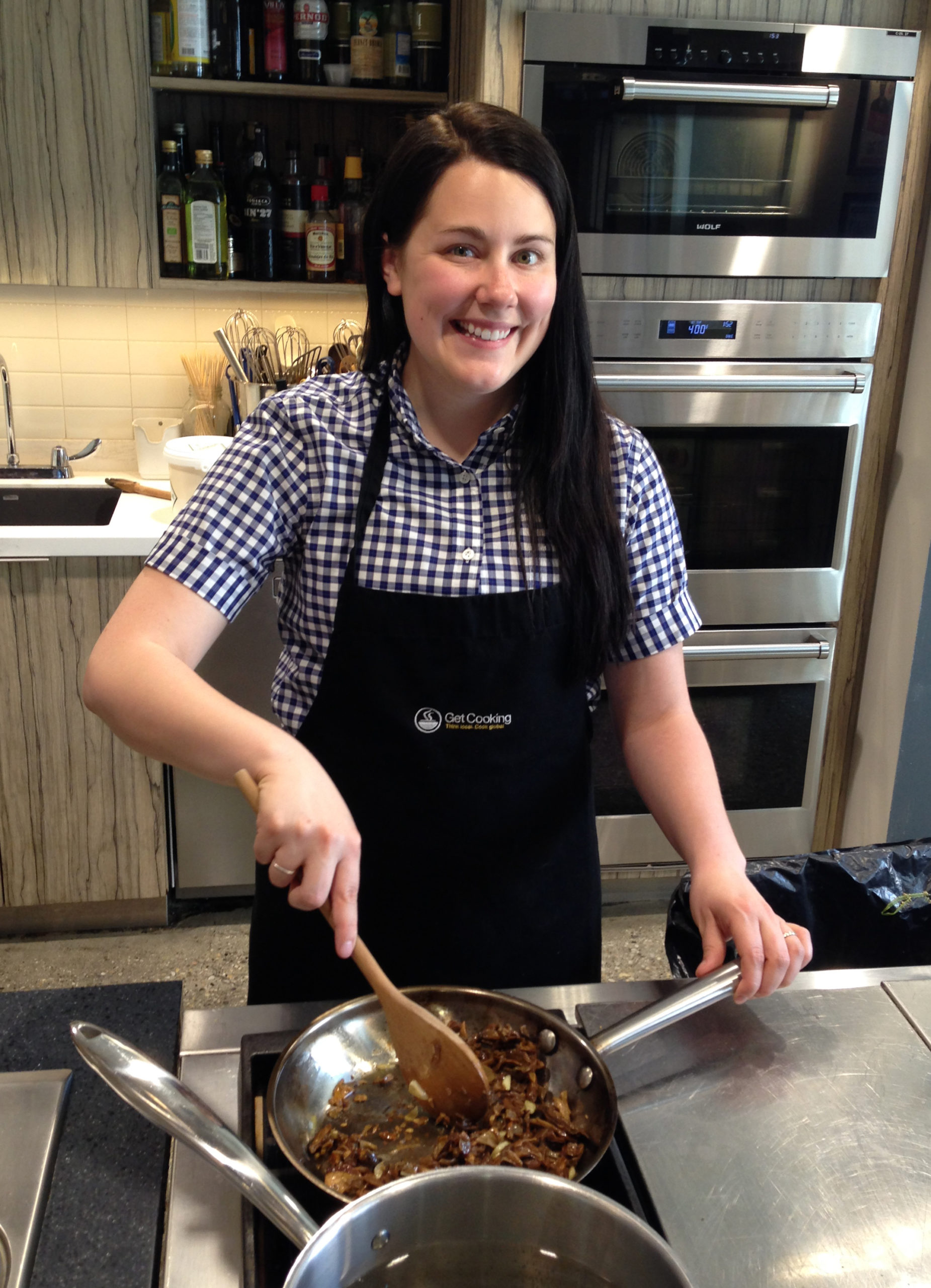 Jen stirs mushrooms in a pan, smiling in a black apron and black and white plaid shirt