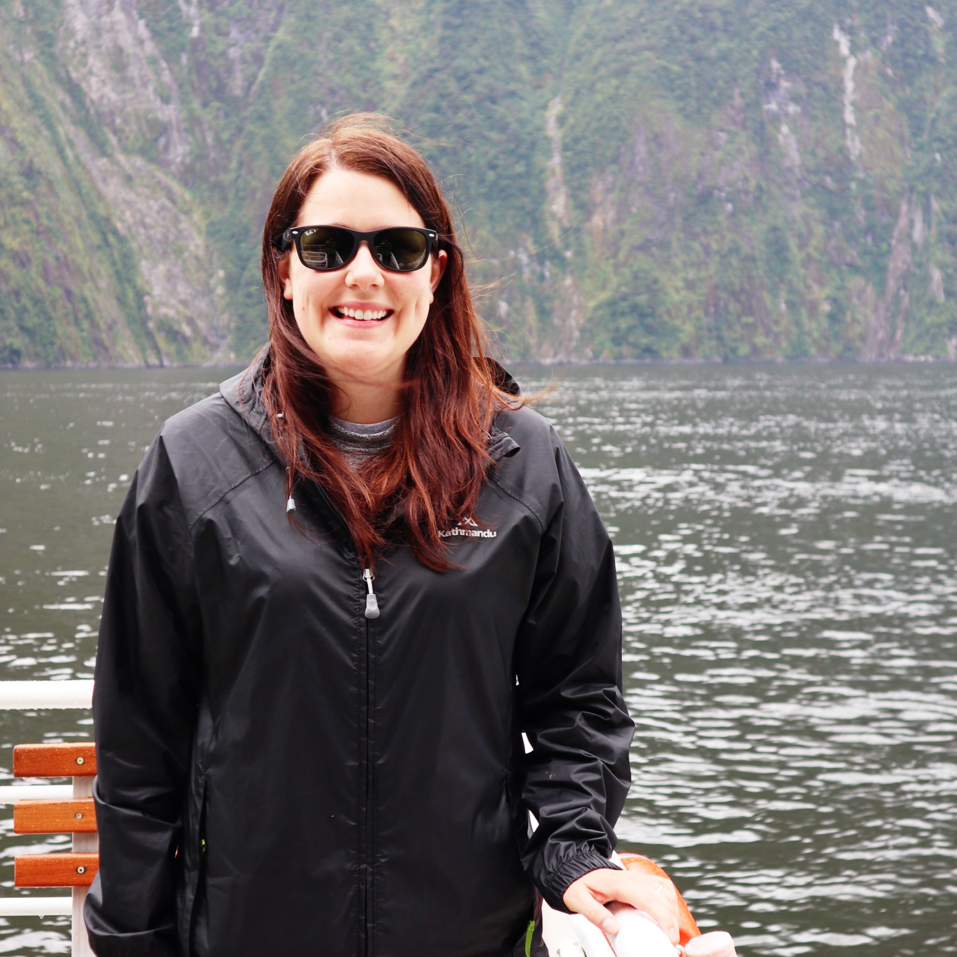 Jen wearing sunglasses and a black rain jacket smiles in front of a lake