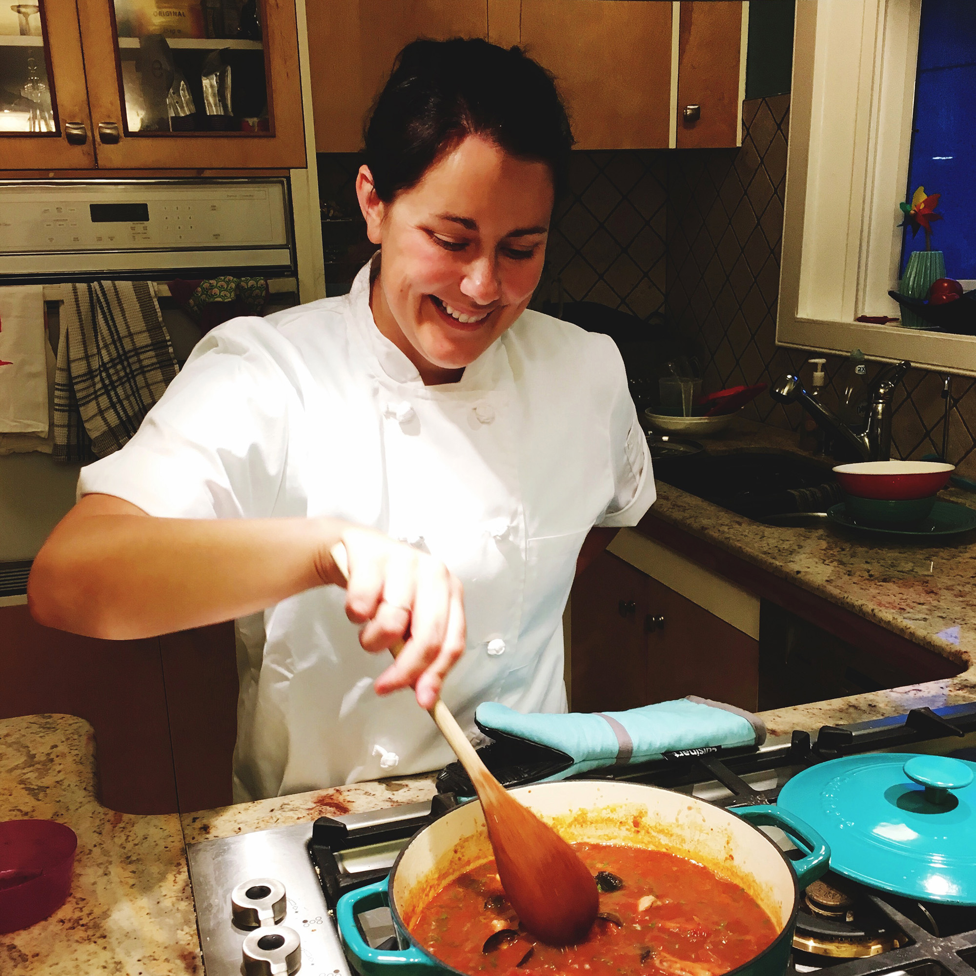 Jen smiles while wearing a chef's jacket and stirring a pot of chili