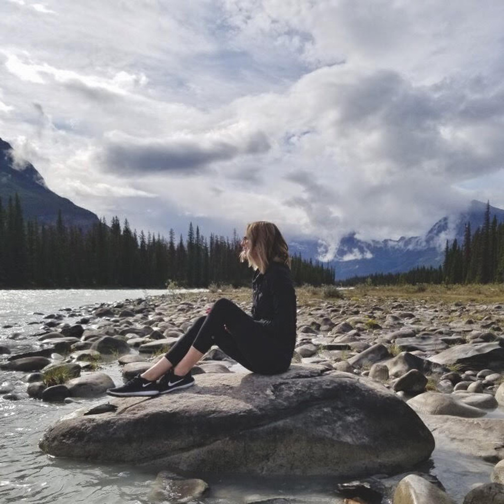Liz sits on a rock in front of a lake and the mountains
