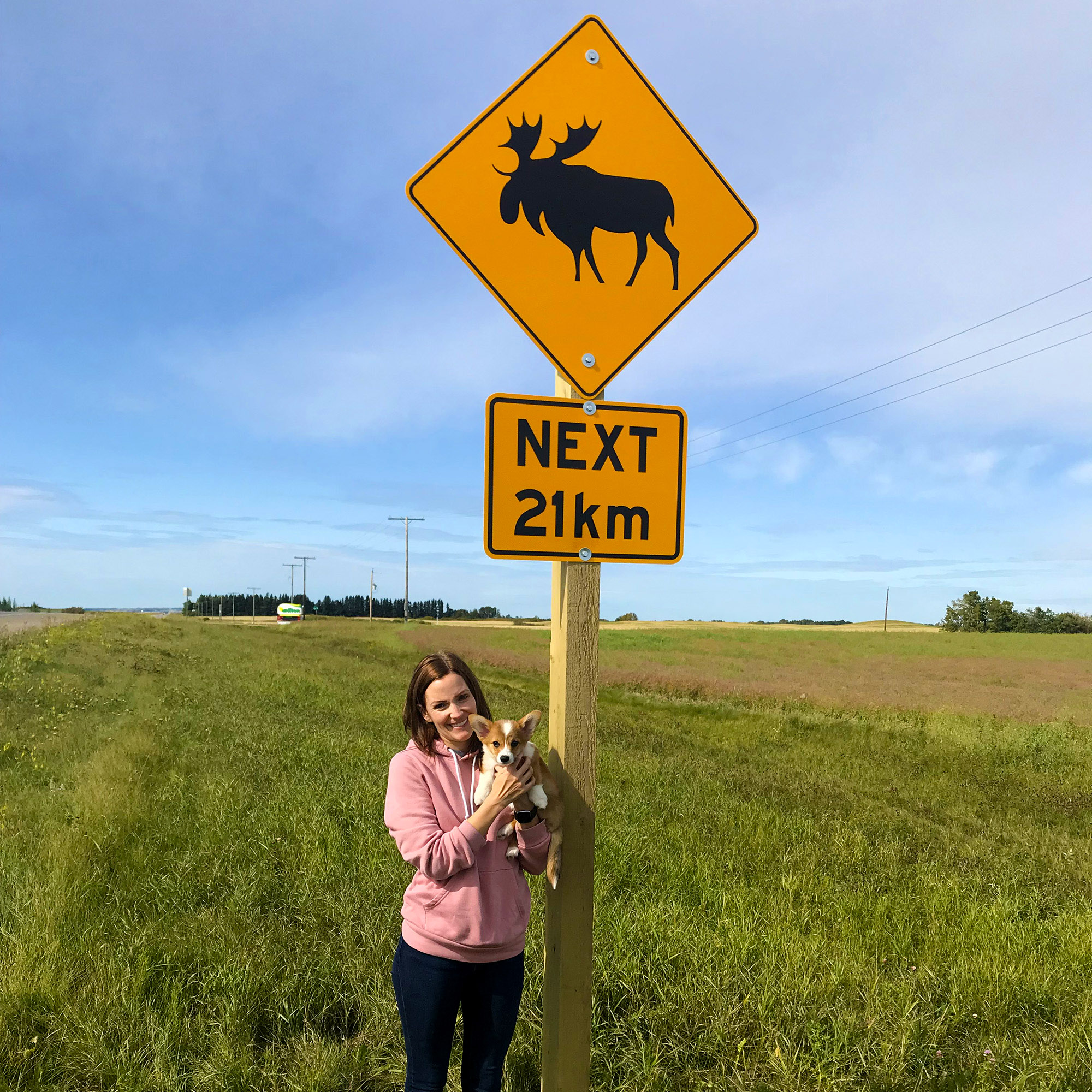 Sarah holds baby Moose, a corgi, in front of a moose crossing sign