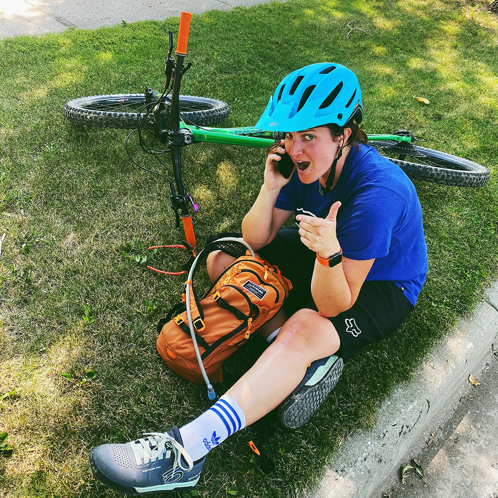 Emma on her phone, giving a thumbs up while sitting on the ground beside her bike