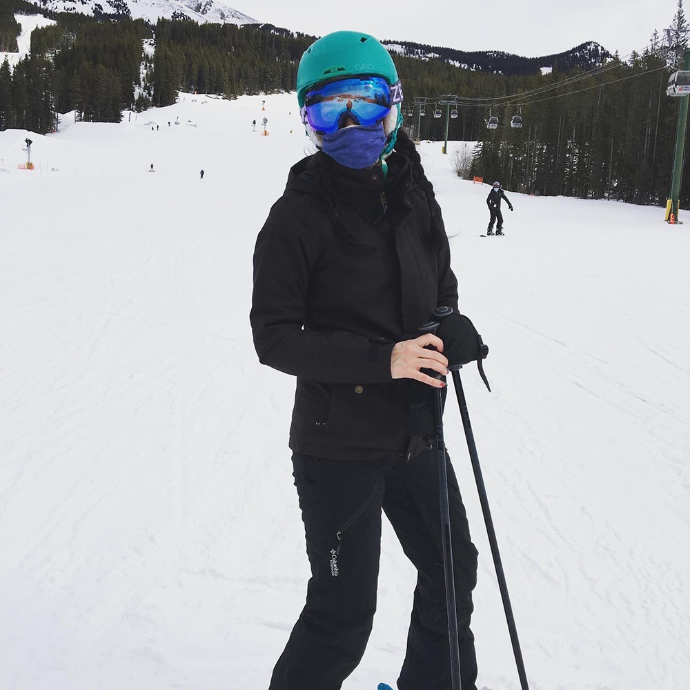 Jessie wears a black ski suit and teal helmet while on a ski hill