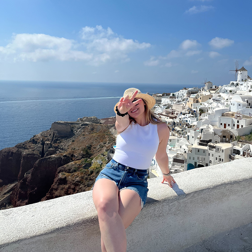 Kyra gives the symbol while sitting on top of a wall looking over Greece and the ocean
