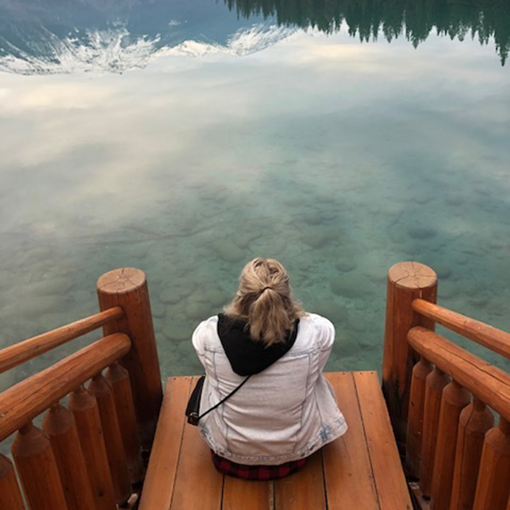 Melenie sits with her back towards the camera. She is sitting on a wood bench looking over a crystal clear lake