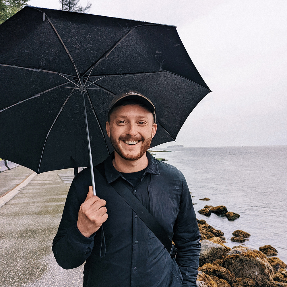 Matt smiles while standing in front of the ocean holding a black umbrella